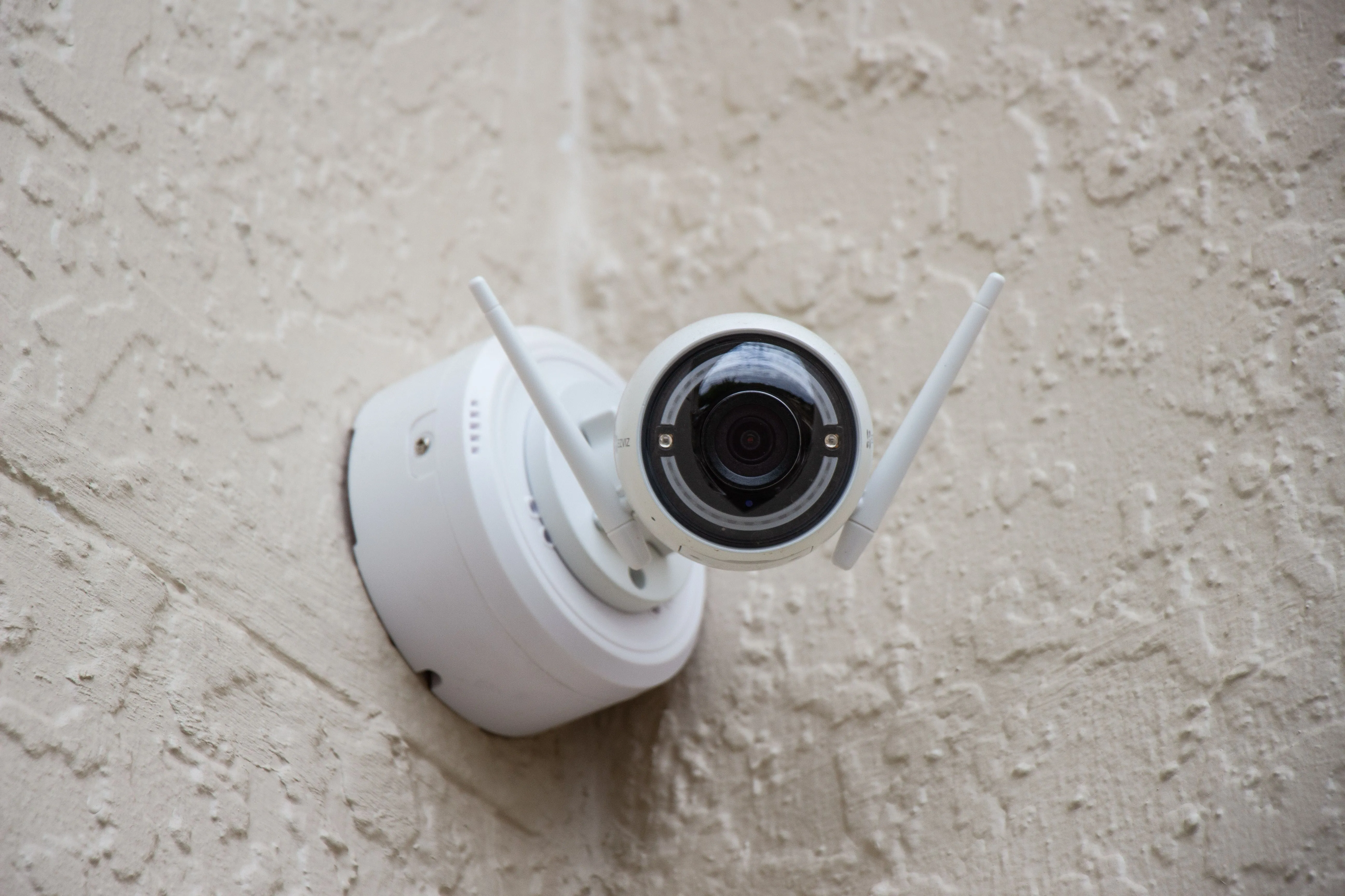 Camera an ninh không dây gắn trên tường màu be, tượng trưng cho hệ thống giám sát gia đình cần truy cập từ xa qua port forwarding.